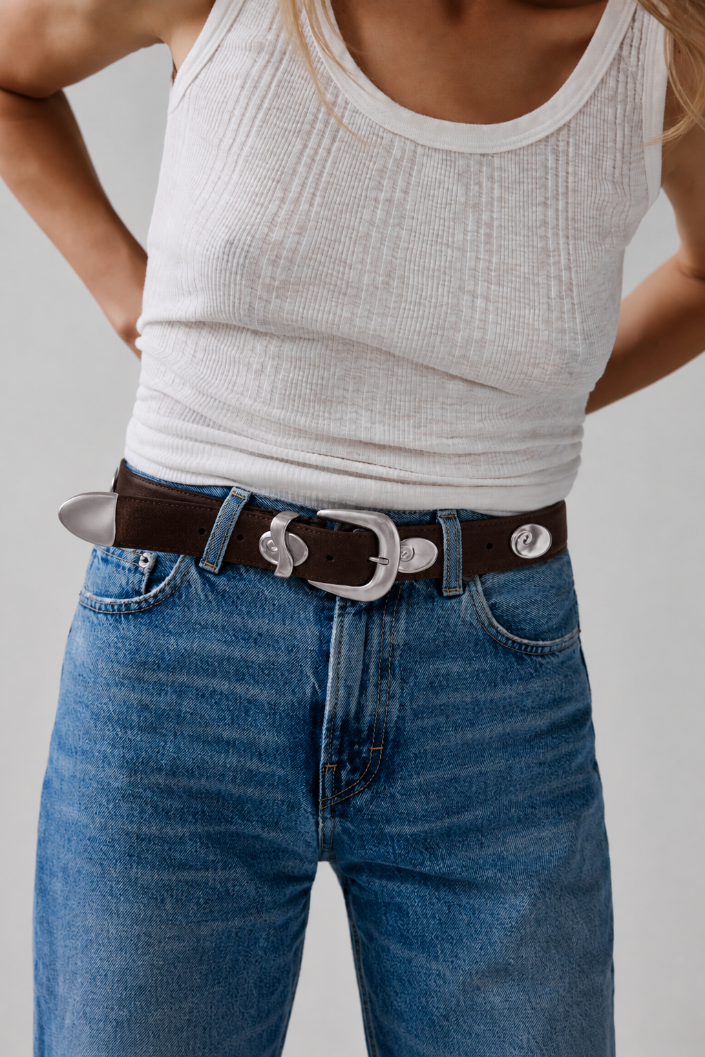 A close-up of a person wearing a dark brown leather belt with a large silver-toned buckle and decorative silver-toned hardware, styled with blue jeans and a white ribbed tank top against a light gray studio background.
